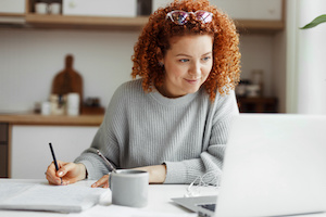 Glad kvinnlig student som studerar online på bärbar dator vid sitt köksbord